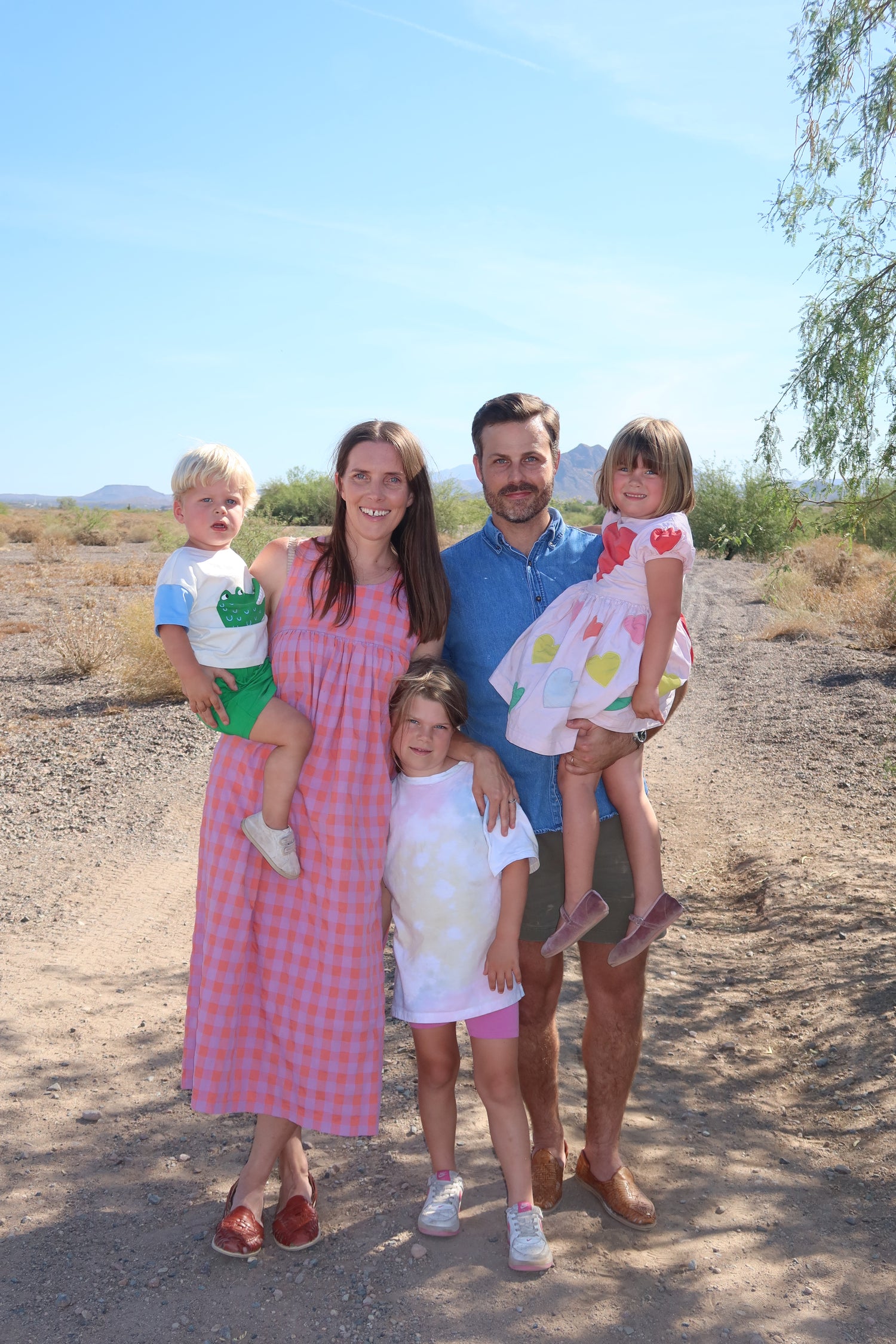 A family of five posing outdoors on a dirt path in a desert landscape under a clear blue sky. The mother wears a pink and white gingham maxi dress, the father wears a blue polo shirt, and the children are dressed in light summer clothing.

Erica Tighe Campbell, Be A Heart